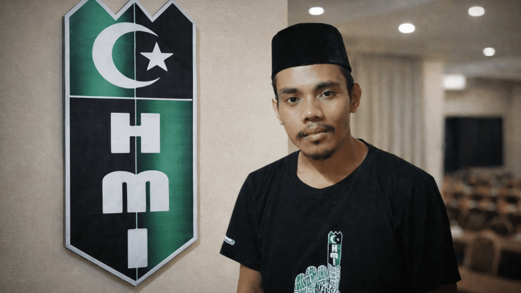 Potret Abdul Salam Ahmad mengenakan peci hitam dan kaos bertema HMI dengan latar belakang simbol bendera Himpunan Mahasiswa Islam.