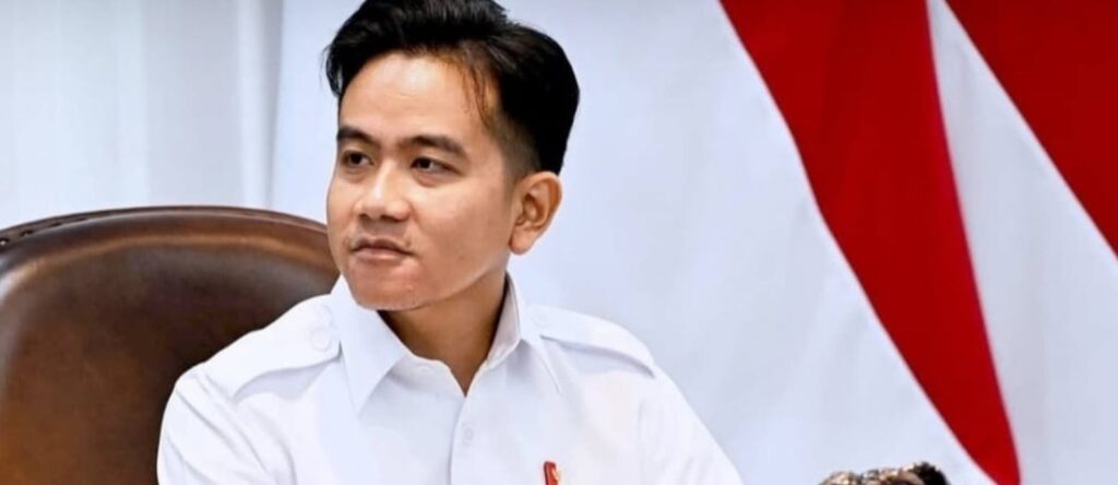 Foto Wakil Presiden Gibran Rakabuming Raka mengenakan kemeja putih formal dengan latar belakang bendera Merah Putih, tampak sedang memberikan pernyataan dengan ekspresi serius.